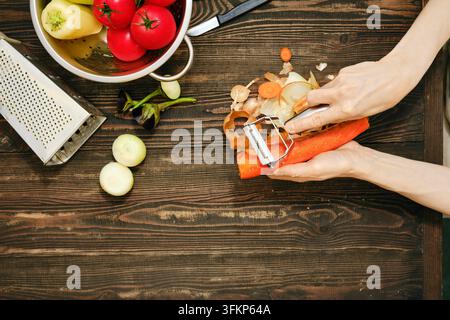 Top view of hands peel carrots while preparing vegetables on wooden table in kitchen. Fresh tomatoes and bell peppers sit in colander Stock Photo
