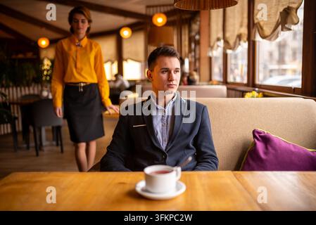 Young man sitting alone at a restaurant table with a woman in a yellow short on background Stock Photo