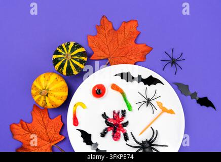 Festive composition with plate of tasty jelly candies and decor of bats, spiders, pumpkins and autumn leaves on purple background for Halloween. Top v Stock Photo