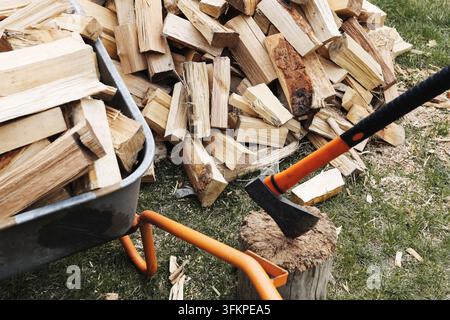 Chopped firewood beside wheelbarrow and splitting axe outdoors. Wood background for heating fuel timber preparation fireplace supplies winter energy Stock Photo