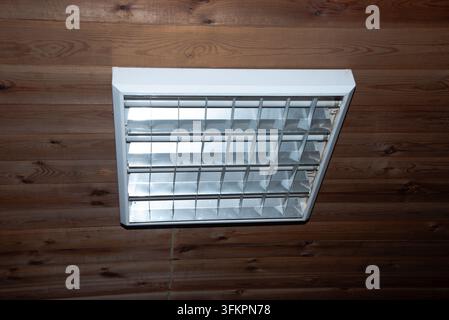 Ceiling-mounted fluorescent light fixture with reflective metal panels on a rustic wooden wall. Stock Photo