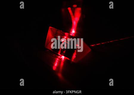 Red laser beams cross a glass prism on a black stage revealing refraction and reflection paths in a moody lab setup that highlights precise optical al Stock Photo