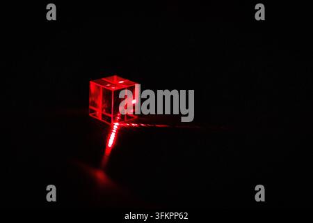 A solitary cube sits in darkness while a red laser enters and exits its faces tracing refracted routes that glow with speckled light and hint at metic Stock Photo