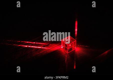 In a deep black lab a cube anchors intersecting red lines and speckled rays that chart a precise optical route ideal for illustrating photonics measur Stock Photo