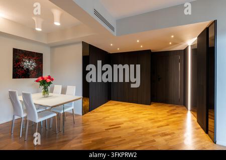 White dining table with pink and red peonies and roses faces a dark mirrored hall across Iroko parquet in a contemporary Italian-designed home. Stock Photo