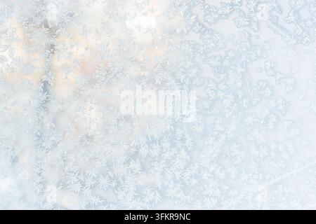 A light background featuring delicate icy frost patterns and snowflakes on window glass with a soft, golden warm glow shining through from behind. Stock Photo