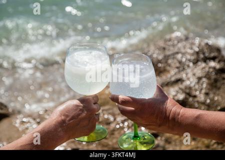 Two people holding glasses of chilled sparkling drinks with ice during a sunny summer day by the sea. Stock Photo