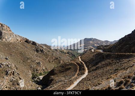 A narrow winding road descending through a rugged rocky gorge toward distant hills and the sea. Stock Photo