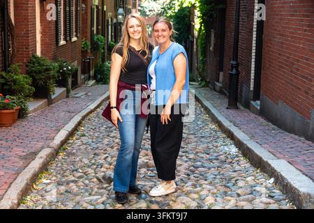 Two women stand close together in quiet narrow cobblestone alley with brick buildings in Boston. Daylight and warm colors highlight their casual outfi Stock Photo