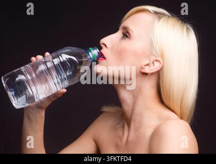Blonde woman tilts head back to drink from a clear plastic water bottle against a dark background, conveying refreshment, hydration, health and simple Stock Photo