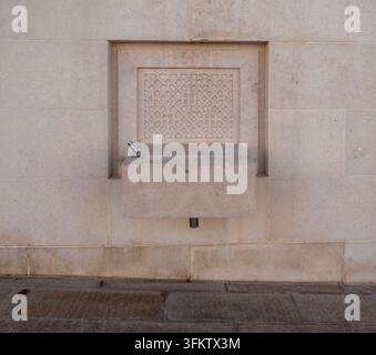 Minimalist outdoor stone drinking fountain with carved geometric Islamic pattern panel mounted on a beige wall in Doha Qatar. Stock Photo