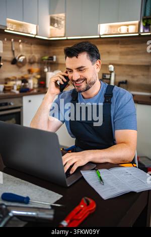 Plumber talking on smartphone and working on laptop in kitchen Stock Photo