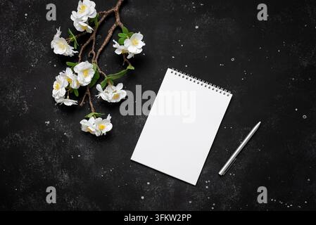 Open blank notepad mockup, pen and decorative cherry blossom branch on black stone background. Spring stationery still life. Top view. Stock Photo