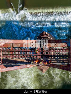 Aerial view of construction workers in high-visibility gear working on a rusted steel railway bridge above a rushing river with white water rapids in Stock Photo