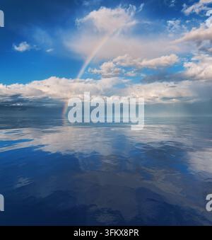 A vibrant rainbow stretches across a calm ocean, framed by scattered clouds and a distant horizon Stock Photo