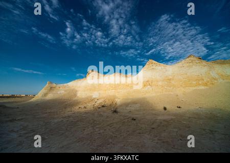 Sunlit white limestone rock formations rise above sandy desert ground under a dramatic deep blue sky filled with textured clouds. Stock Photo