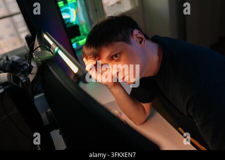 Concentrated young man with thoughtful expression works or plays on computer at night. Angled shot shows intense focus under monitor light. Modern wor Stock Photo