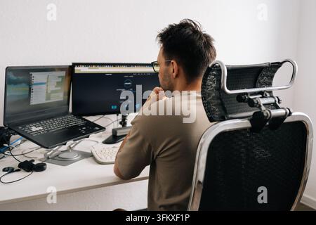 Rear shoulder view of young man developer focused on work at his desk. Modern home office setup with laptop and external monitor. Concept of freelance Stock Photo