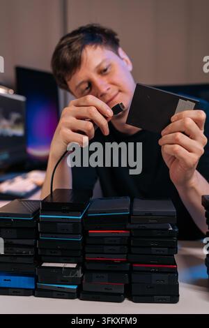 Focused technician connecting portable hard drive. Many external drives stacked on desk represent big data, information backup, storage management, or Stock Photo