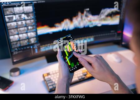 Person hands holding smartphone displaying candlestick chart, analyzing financial market trends and investment analytics with computer monitor showing Stock Photo