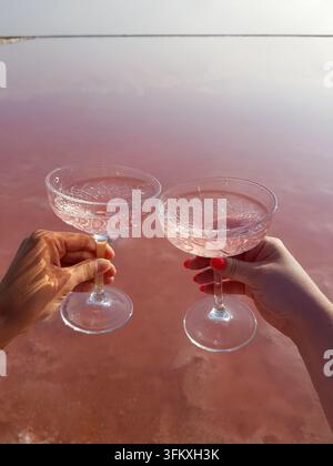 Point of view shot of two women hands toast crystal glasses filled with rose wine or cocktail with natural salt lake background on a sunny day. Stock Photo