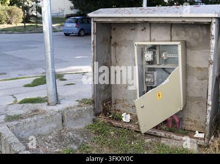 Broken metal electrical cabinet standing open on a sidewalk in a city, revealing exposed wiring and circuit breakers, representing neglect, vandalism, Stock Photo