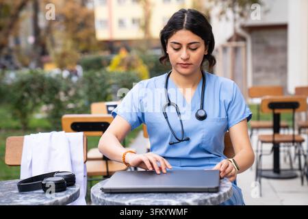 Young female healthcare professional preparing a laptop for an online consultation while wearing blue medical scrubs and a stethoscope. Ideal for tele Stock Photo