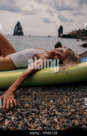 Blonde woman lying comfortably on paddleboard at pebble beach during overcast day Stock Photo