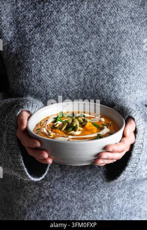 Female hands in gray knitted sweater holding bowl of pumpkin soup close up. Autumn cozy meal, seasonal comfort food concept. Healthy eating. Stock Photo