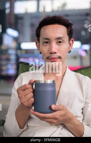 A high quality professional vertical lifestyle portrait of a confident Thai non-binary gay man with an outdoor drink. The model looks at the camera le Stock Photo