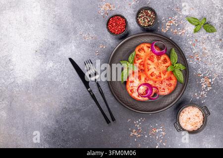 Red tomatoes on a black platter, with purple onion rings and green basil leaves, accompanied by black cutlery and scattered spices on a gray concrete Stock Photo