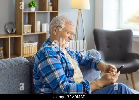 Happy senior man using smartphone at home on sofa, smiling and relaxed indoors Stock Photo