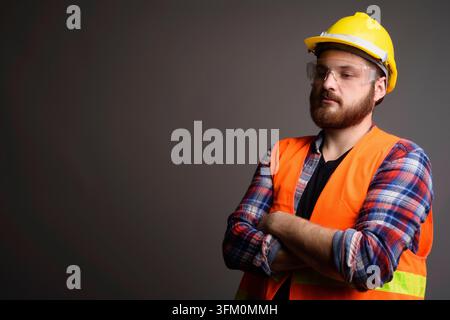 Studio portrait of a serious bearded man in an orange safety vest and yellow hard hat with folded arms down. features large copy space for engineering Stock Photo