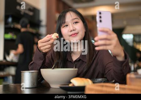 A horizontal photograph of a young Asian woman looking at her pink smartphone held up in her left hand to take a selfie or record a video. She holds a Stock Photo