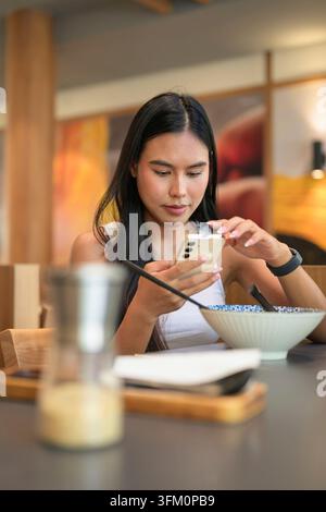 Vertical medium shot of a focused young Isan Thai female client with a serious, focused expression while her smartphone with both hands. She sits behi Stock Photo