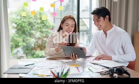 A young Asian man and woman sitting at a desk, smiling while reviewing mobile application wireframes on a digital tablet in a bright modern workspace Stock Photo