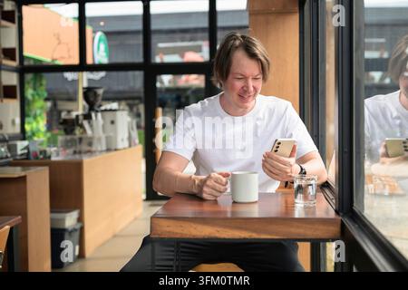 A happy, smiling Caucasian Scandinavian man in his late 30s or early 40s sits on a wooden table next to a large glass window in a modern coffee shop. Stock Photo
