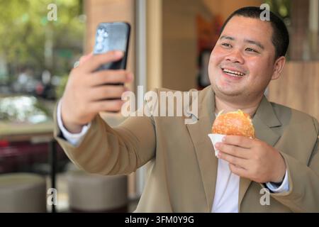 A happy plus size mid adult Asian businessman from the Isan region takes a smartphone selfie while holding a hamburger at a restaurant table with a sh Stock Photo