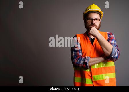 Studio portrait of a thoughtful bearded man in an orange safety vest and yellow hard hat touching his chin. Features large copy space for industrial, Stock Photo