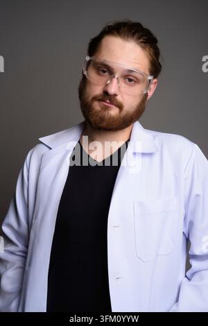 Studio portrait of a bearded young man in a white lab coat and protective safety goggles looking at the camera with a serious expression. Concept for Stock Photo
