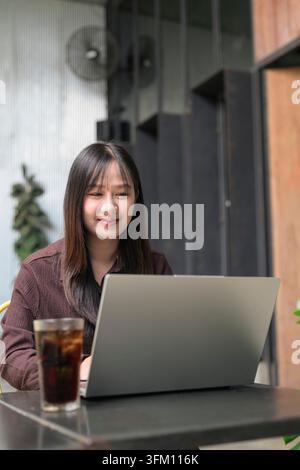 A vertical, medium close-up photograph of a young Asian woman looking at her silver laptop with a soft, satisfied smile. She is seated at an outdoor p Stock Photo