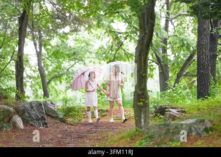 Two women are walking in a forest, holding umbrellas to protect themselves from the rain. The umbrellas are pink and white, and the women are dressed Stock Photo