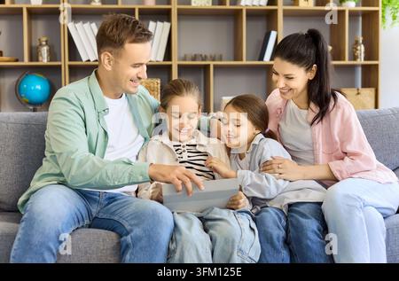Cheerful family parents with daughters using tablet on sofa at home, online fun Stock Photo