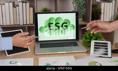 Two people point at a laptop screen displaying an ESG report, with a small solar panel on the desk, symbolizing sustainable business practices Stock Photo