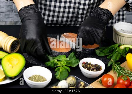 Hands preparing salmon steak for cooking, next to table basil, rosemary spices and bell peppers, tomatoes, avocado and quail eggs all placed on black Stock Photo