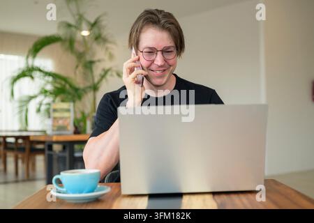 Scandinavian man sits on a wooden table in a bright cafe, smiling while holding his smartphone to his ear and looking at his laptop. He is an adult or Stock Photo