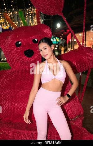 A confident young Asian woman wearing a pink fitness outfit poses with her hand on her hip near a large red teddy bear at night Stock Photo
