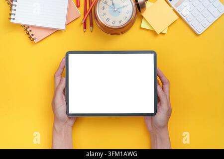 A tablet held by hands surrounded by school supplies on a yellow background, symbolizing digital learning and back-to-school preparation Stock Photo