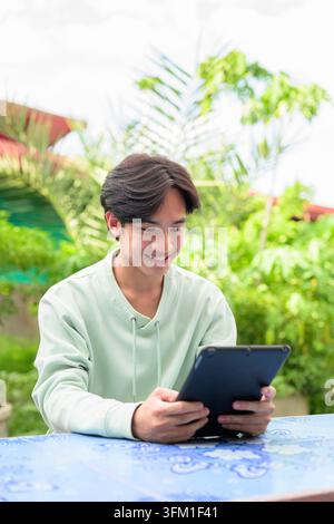 A vertical lifestyle portrait of a cheerful young Asian student or remote worker from the Isan region of Thailand sitting outdoors at a blue tiled tab Stock Photo