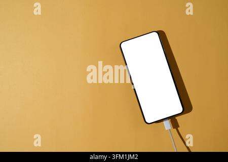 An overhead flat lay of a smartphone with a blank white screen plugged into a white charging cable on a solid yellow background Stock Photo
